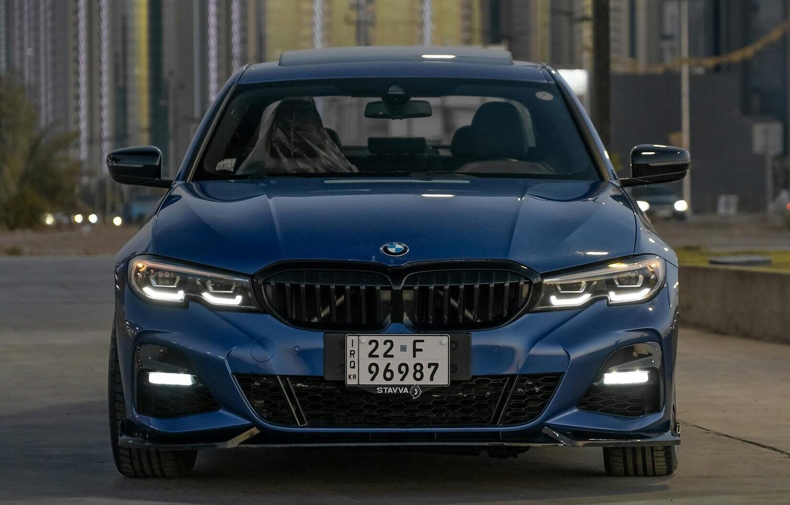 Front view of a blue BMW 3 Series parked on an urban street with city backdrop.